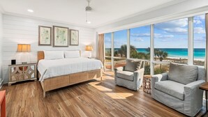 Ein Schlafzimmer mit einem großen Bett, zwei Nachttischen, einem Schminktisch, einem Fenster mit Blick auf den Strand und einer Sitzecke mit zwei Sesseln.