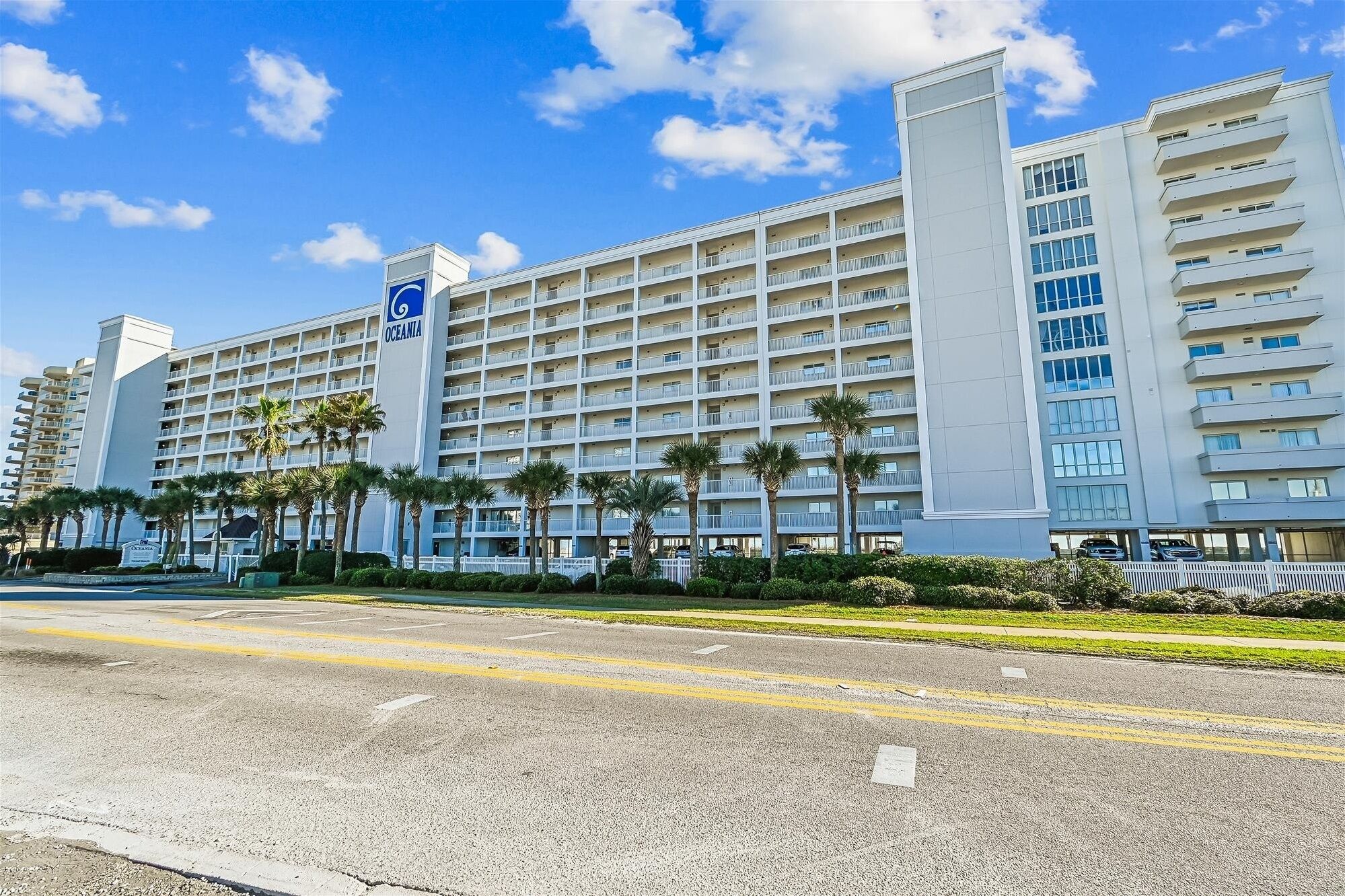 Ein großes, mehrstöckiges Hotel mit einem gut sichtbaren Oceania-Schild, das ein modernes architektonisches Design mit Balkonen und einem Parkplatz davor aufweist.