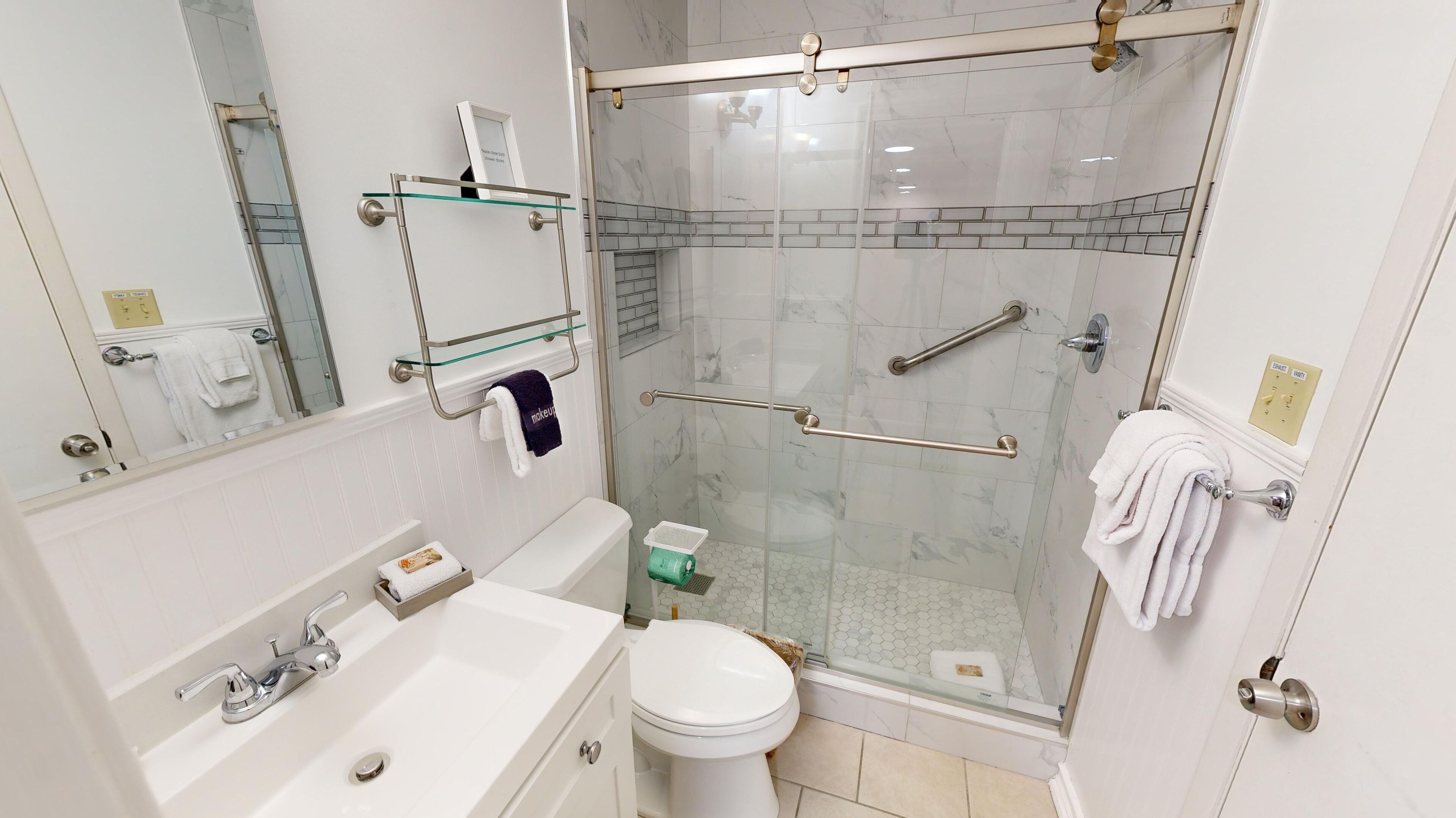 A modern bathroom with a glass-enclosed shower, a white sink, a toilet, and a glass shelf above the sink.