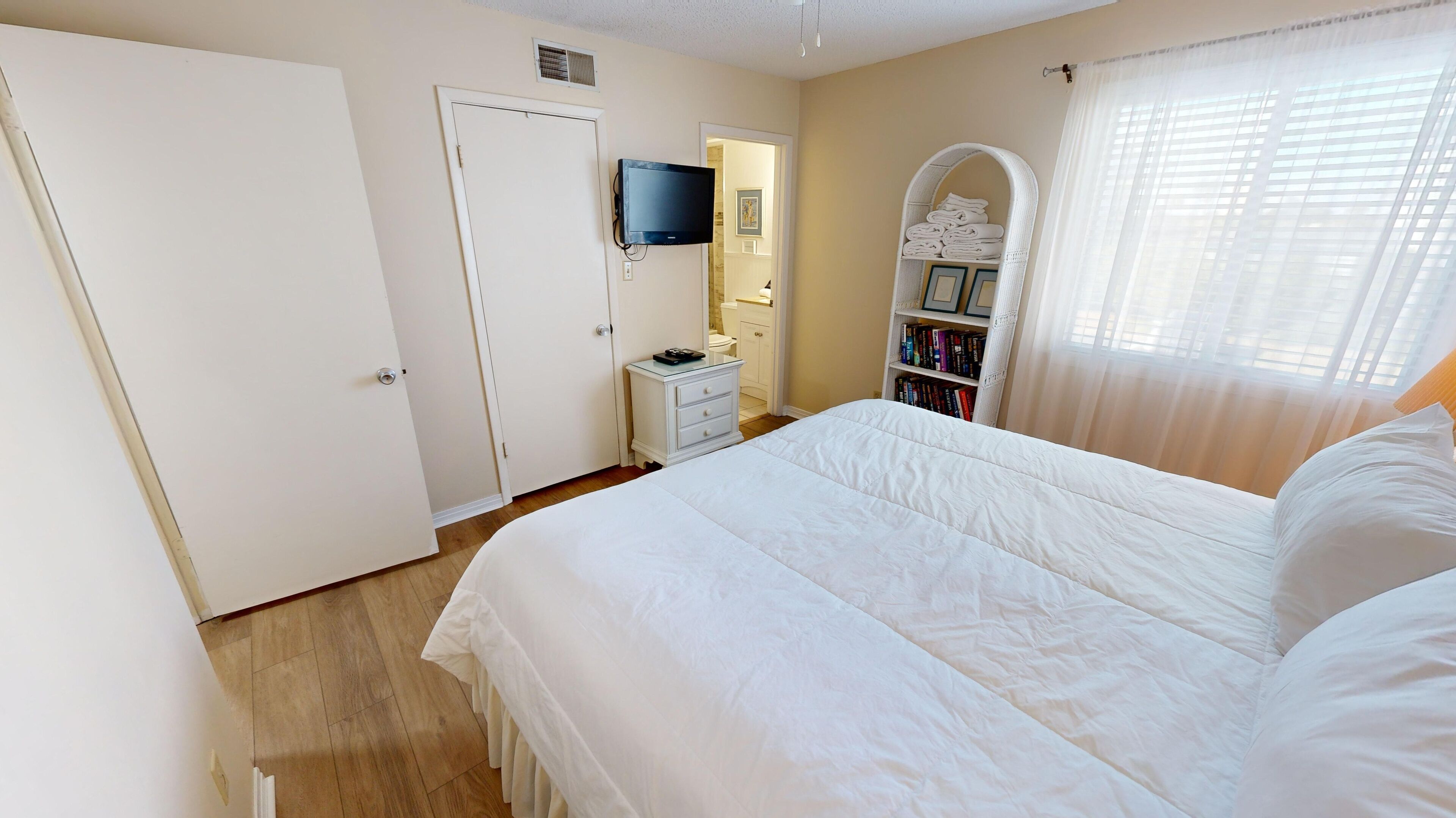 Une chambre avec un grand lit, un téléviseur fixé au mur, une table de chevet, une bibliothèque et une salle de bain visible à travers une porte ouverte.
