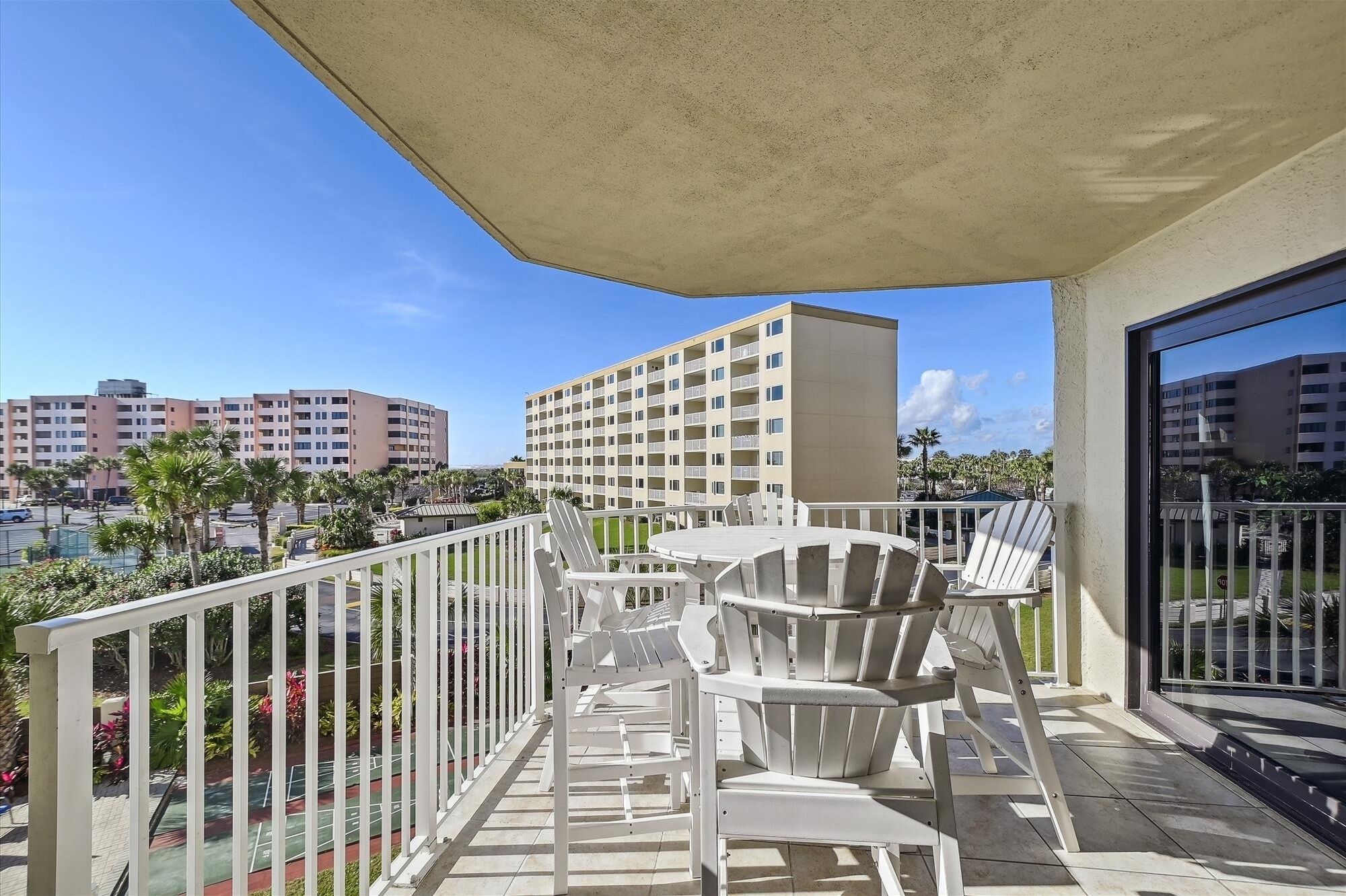 A balcony with a table and chairs, a view of palm trees, and a building in the background.