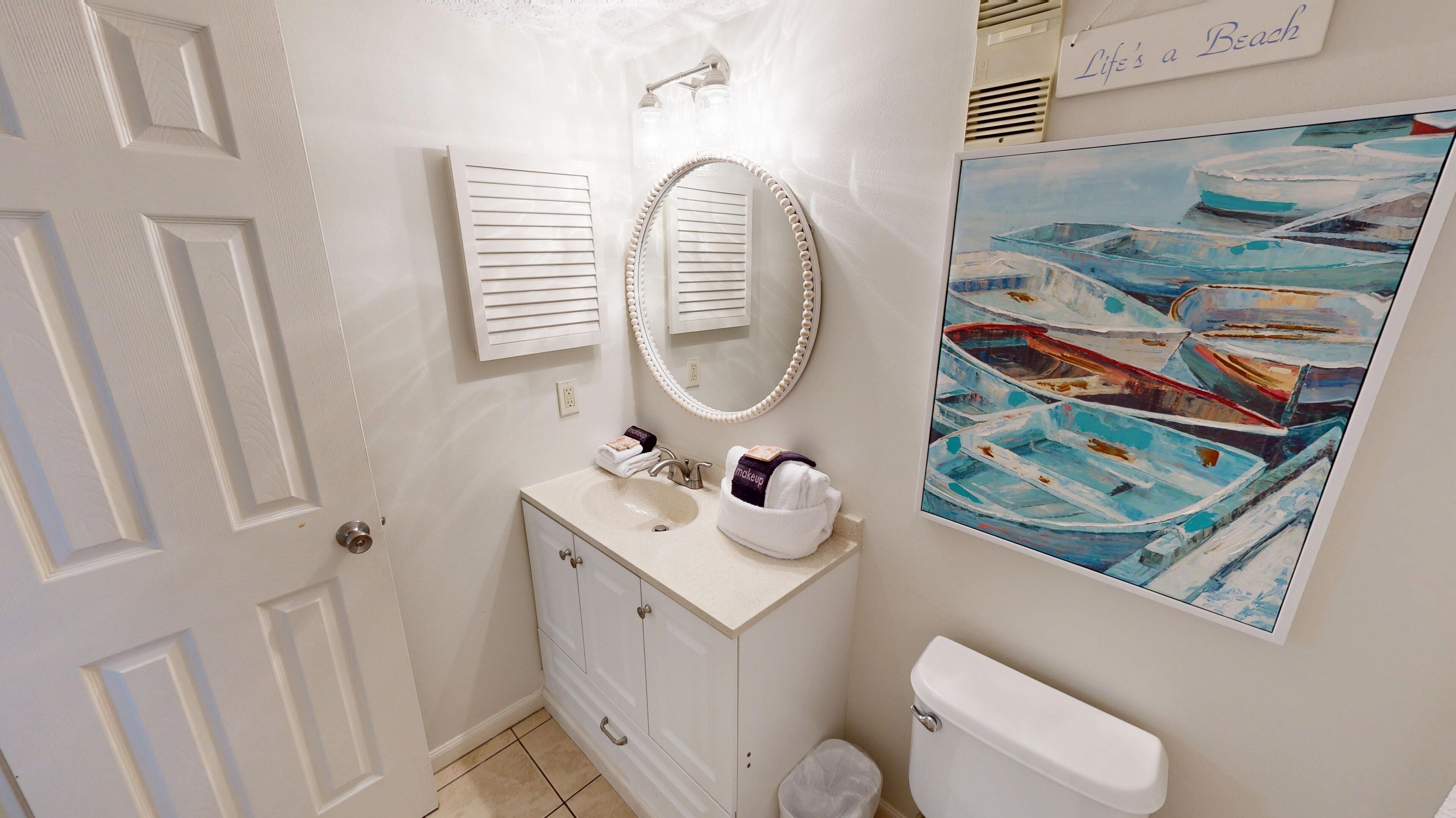 A bathroom with a white vanity, a mirror, a sink, and a toilet. There is a painting of boats on the wall.