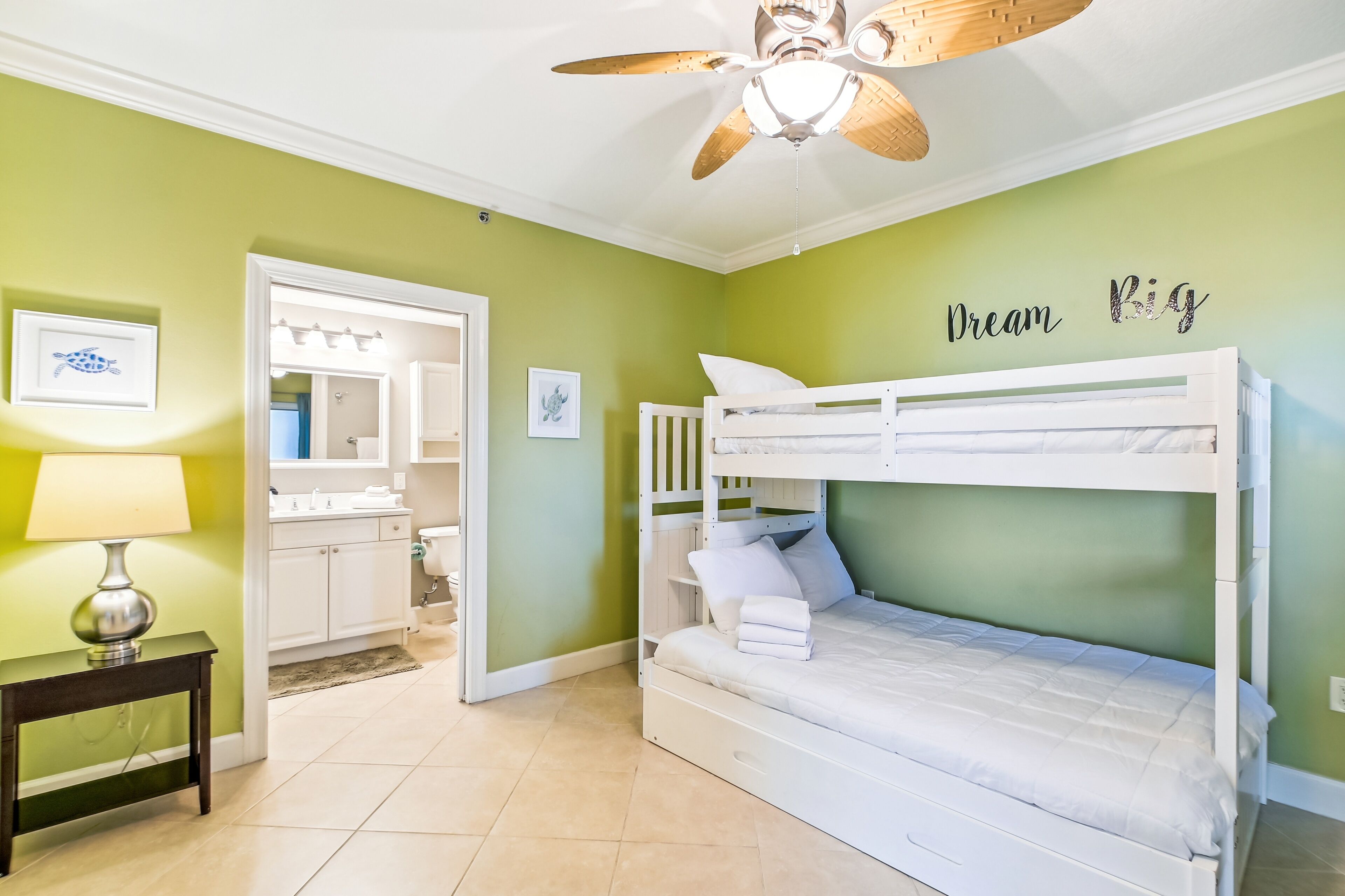 Un dormitorio con literas, ventilador de techo, mesita de noche con lámpara y baño visible a través de una puerta abierta.