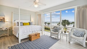 Un dormitorio con un ventanal grande, una cama, un aparador y una silla de mimbre.
