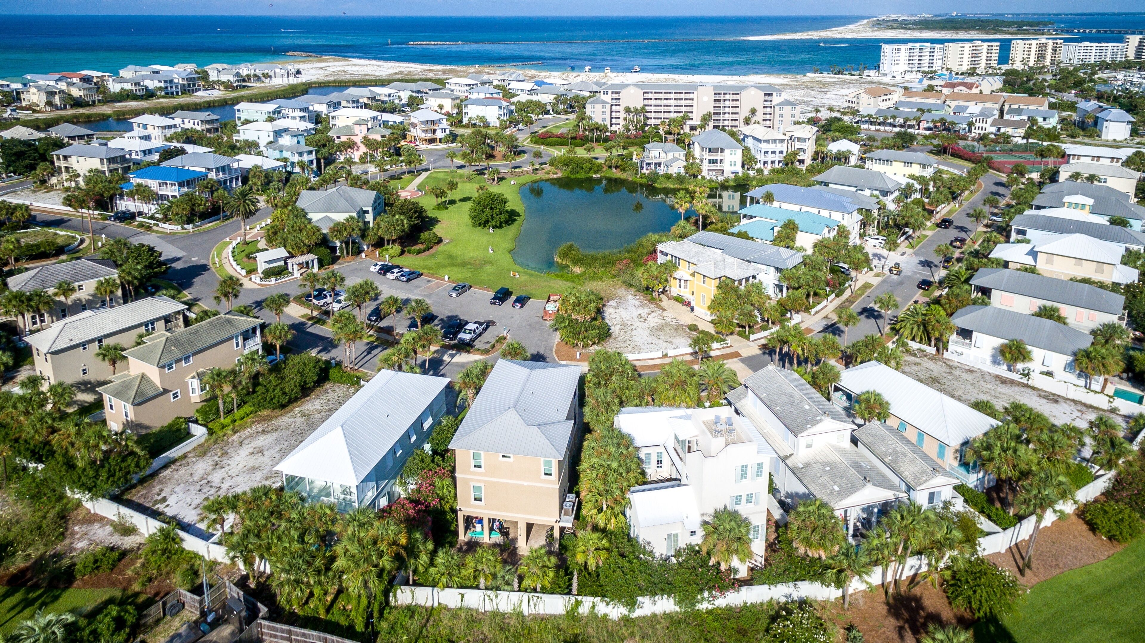 Vista aérea de una zona residencial costera con un estanque central, rodeada de casas de diversos colores y estilos de techos, y una playa al fondo.