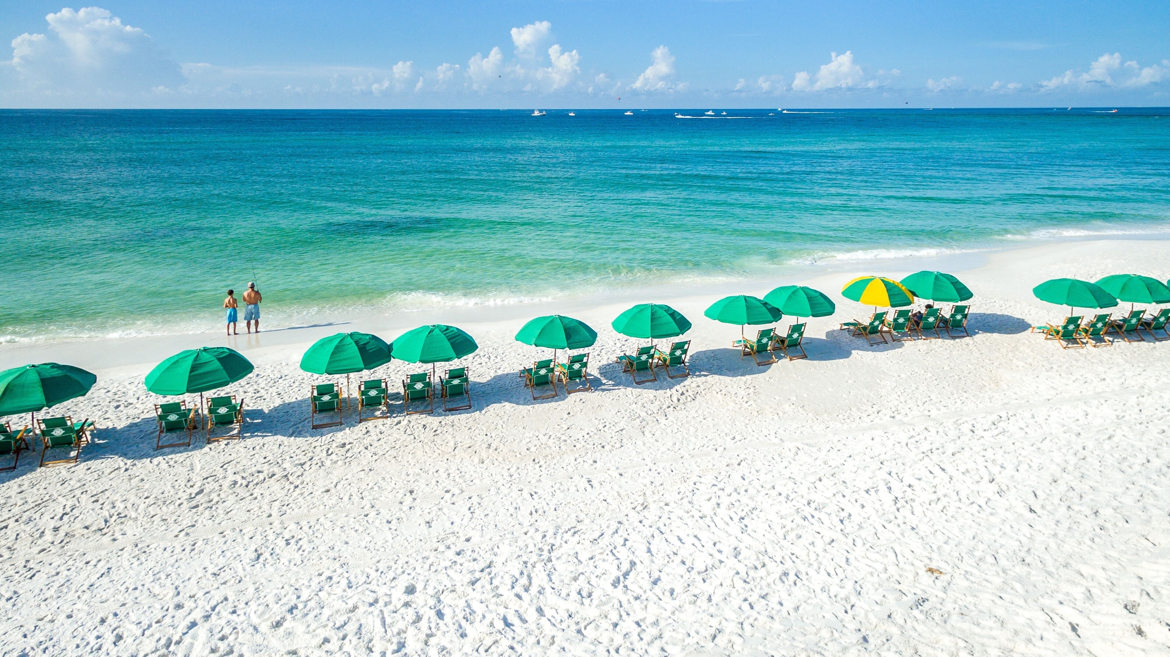 A beach with white sand and turquoise water. There are rows of green umbrellas and lounge chairs along the shore.
