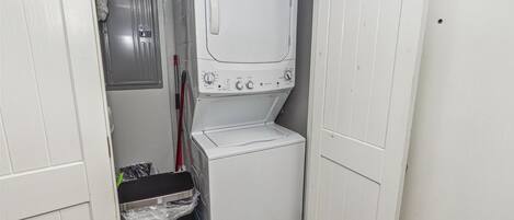 Une petite buanderie équipée d’un lave-linge et d’un sèche-linge superposés, d’une poubelle et d’un balai.