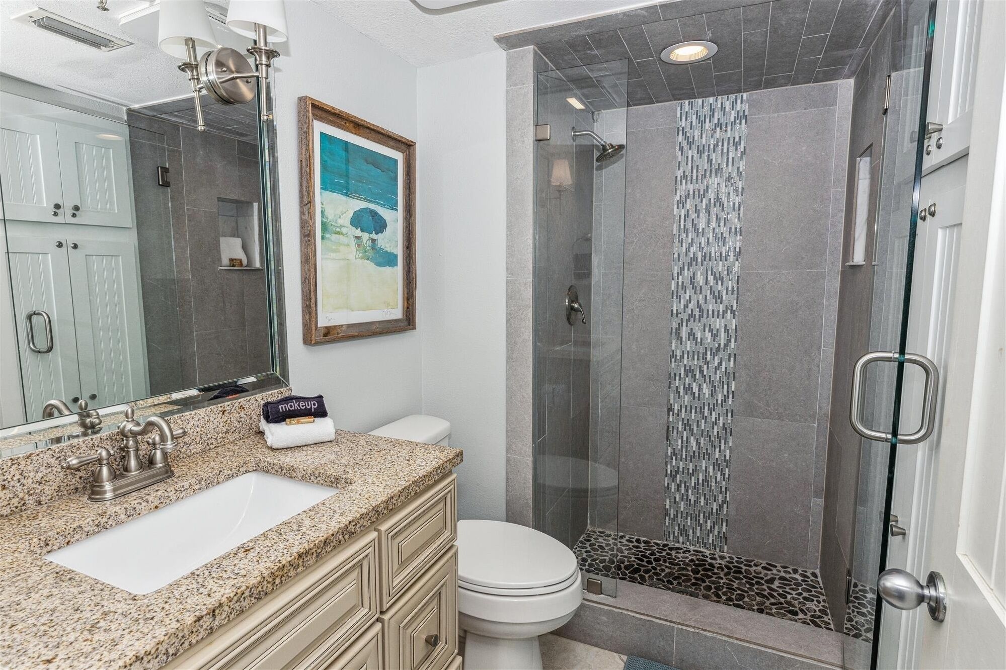 A bathroom with a granite countertop, a white sink, a toilet, and a glass-enclosed shower area with a mosaic tile wall.