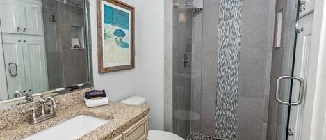 A bathroom with a granite countertop, a white sink, a toilet, and a glass-enclosed shower area with a mosaic tile wall.