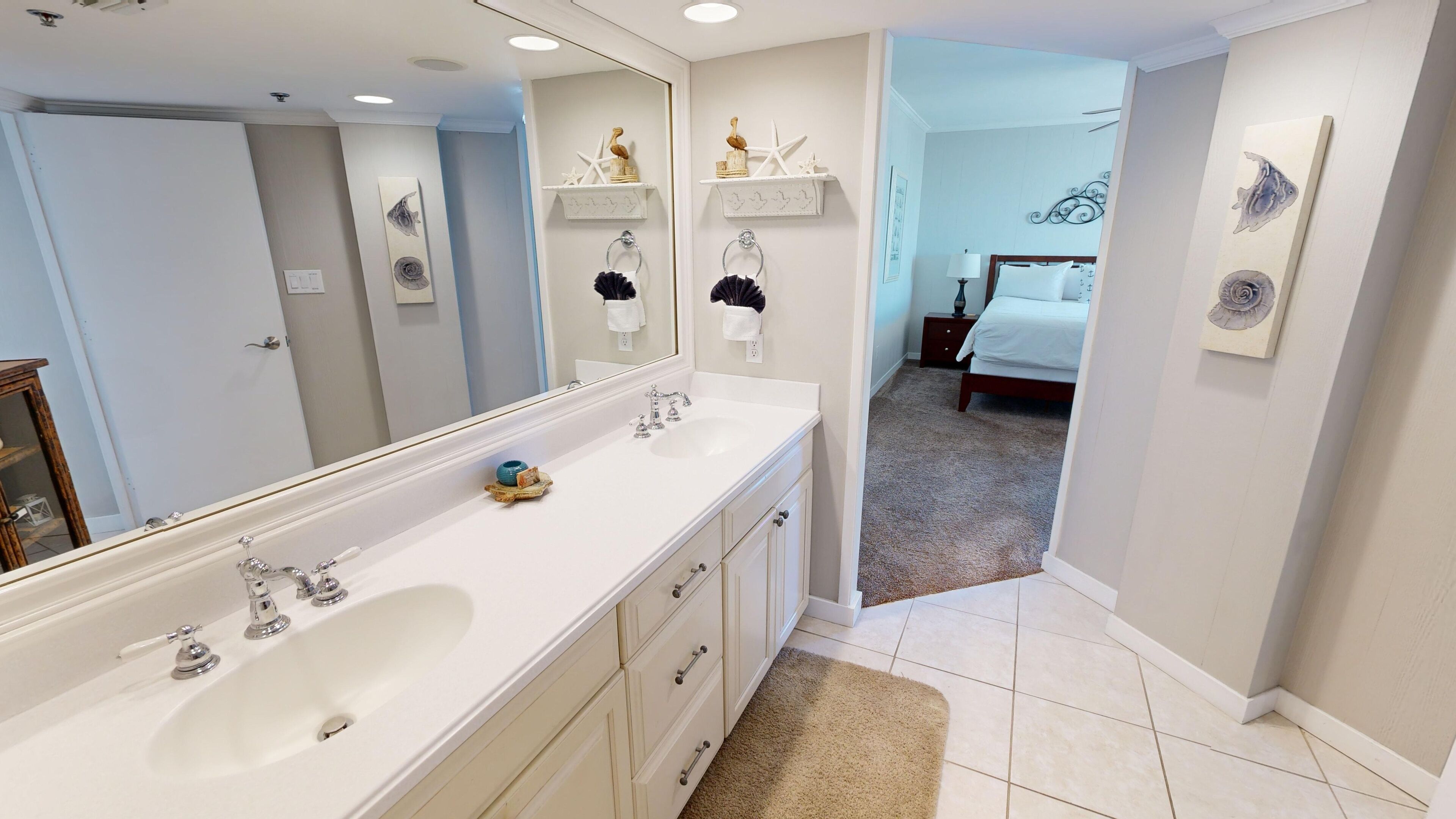 A modern bathroom with a large mirror, dual sinks, and a vanity. There is a hallway leading to a bedroom with a bed and a nightstand.