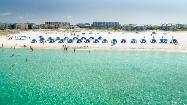 Una playa con agua turquesa cristalina, numerosos sombrillas azules y una hilera de edificios al fondo.