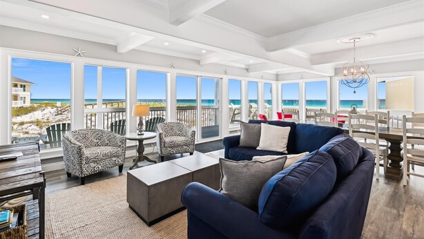 A spacious living room with a large window overlooking the beach, a dark blue sofa, patterned armchairs, and a dining area with a chandelier.