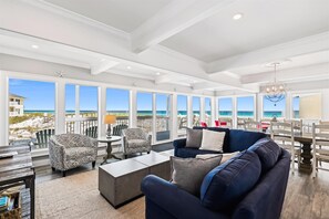 Un vaste salon doté d’une grande fenêtre donnant sur la plage, d’un canapé bleu foncé, de fauteuils à motifs et d’un coin repas agrémenté d’un lustre.