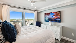 A bedroom with a large bed, a ceiling fan, a TV, and a view of the beach.