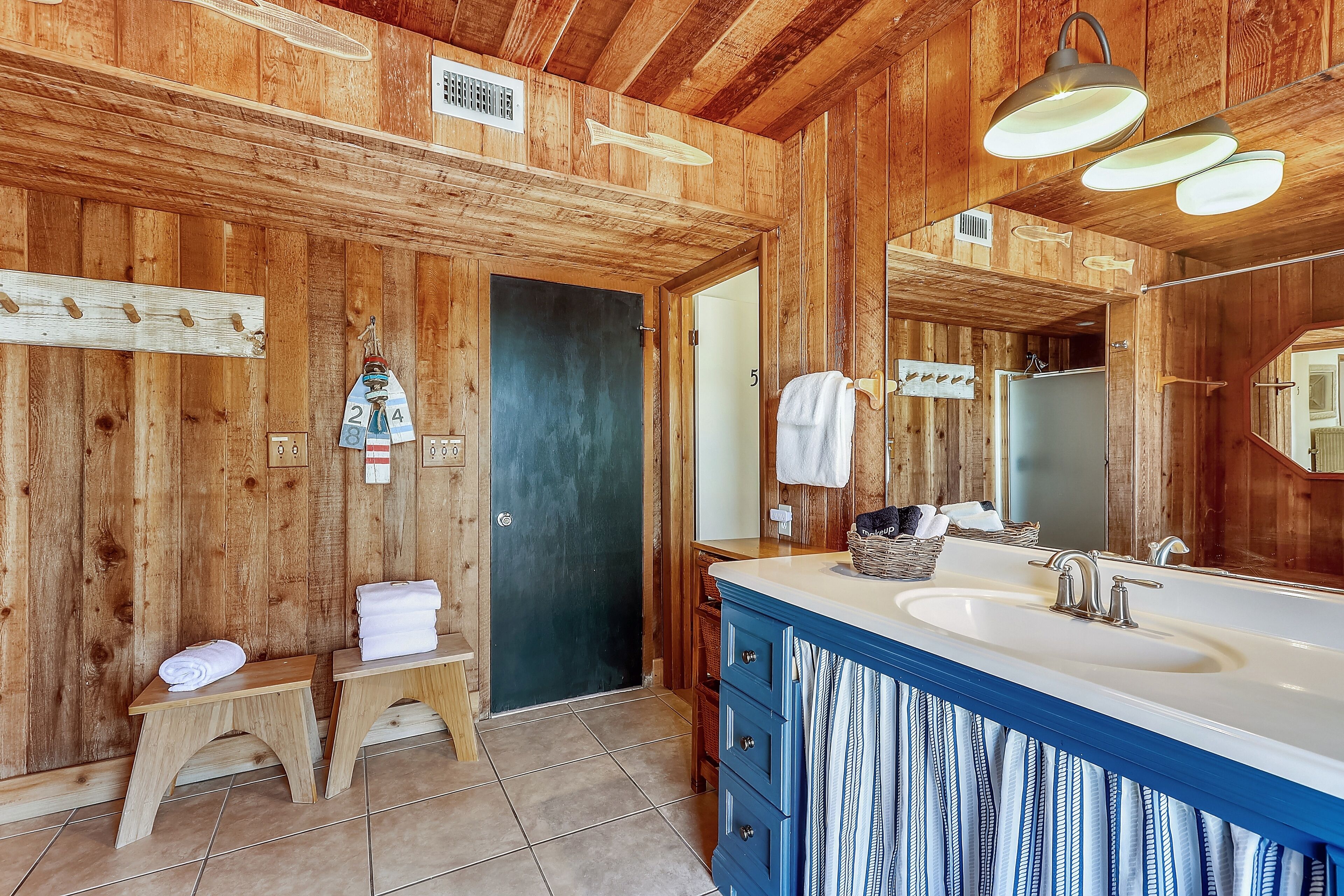 A wooden bathroom with a large mirror, a sink, and two small stools with towels.