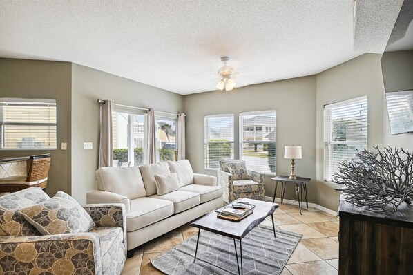 A spacious living room with a beige sofa, a patterned armchair, a coffee table, and a decorative plant. - Sandpiper Cove 9125 2 Bedroom Condo by RedAwning (Destin)