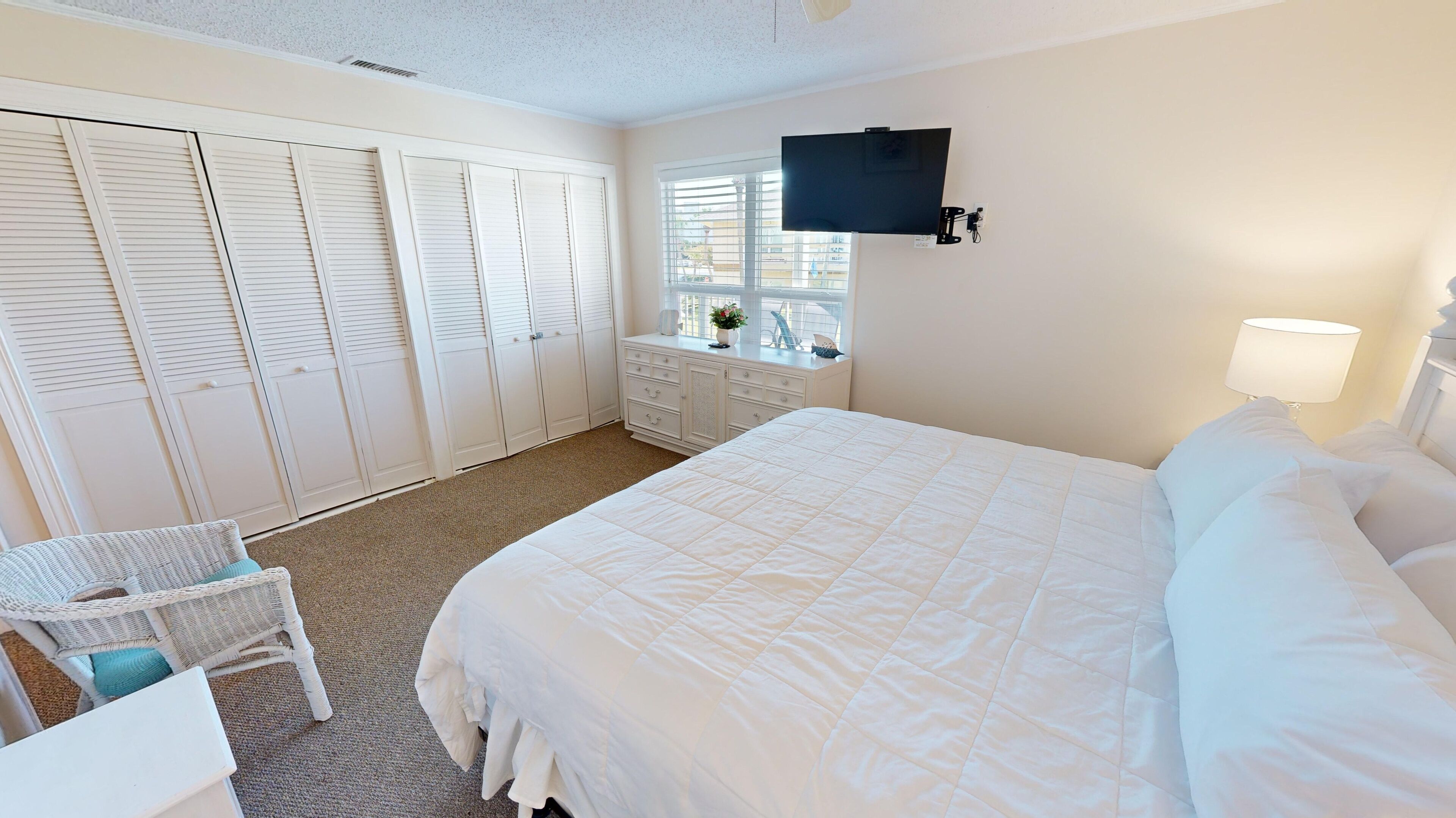 A bedroom with a large bed, a desk with a chair, a TV mounted on the wall, and a window with blinds.