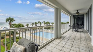 Uma varanda com vista para uma piscina e a praia, área de refeições com mobiliário de exterior e ventoinha de teto.