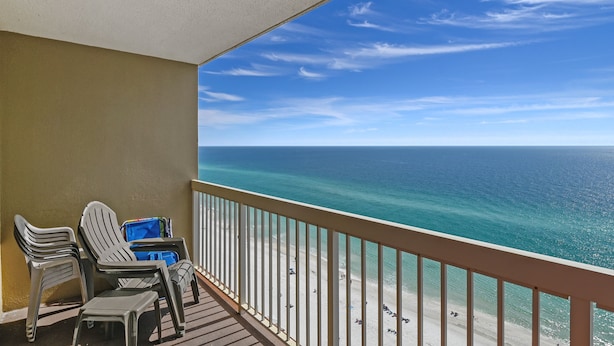 A balcony with two white plastic chairs, a small table, and a blue bag. The balcony overlooks a beach with clear blue water and a sandy shore. - Pelican Beach 1811 1 Bedroom Condo by RedAwning (Destin)