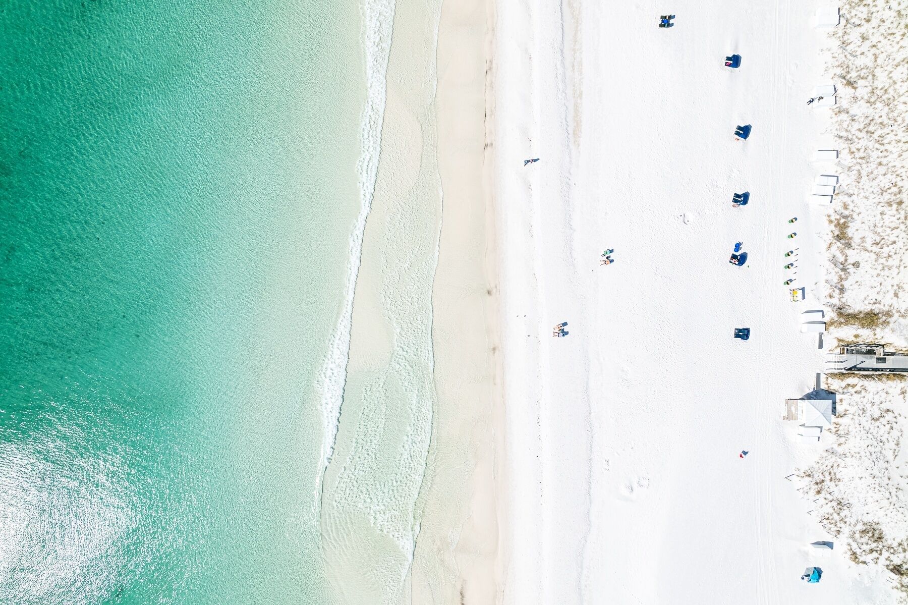 An aerial view of a beach with clear turquoise waters, white sand, and a few people and umbrellas.
