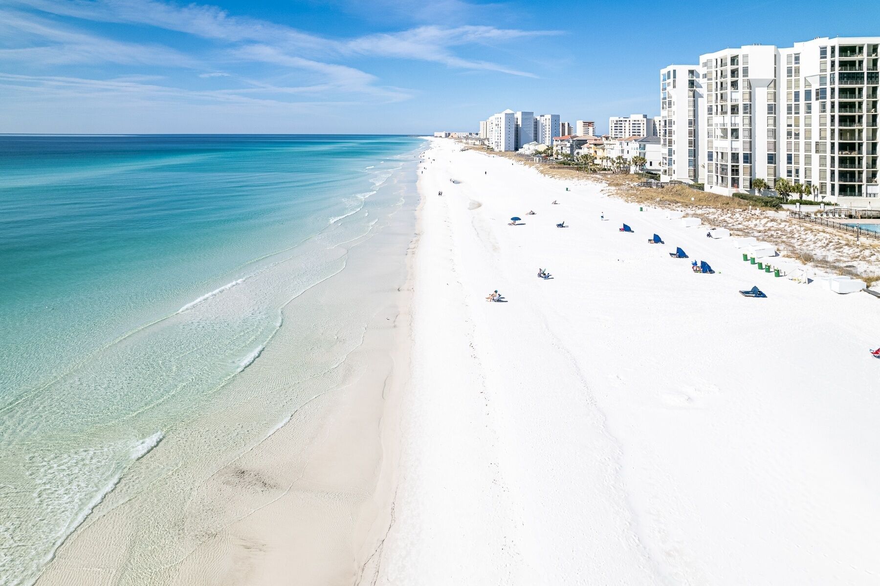 A beach with white sand and clear turquoise water. There are several buildings along the shore.