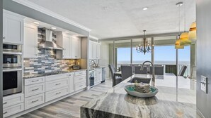 A modern kitchen with white cabinetry, a marble island, and a view of the ocean through large windows.