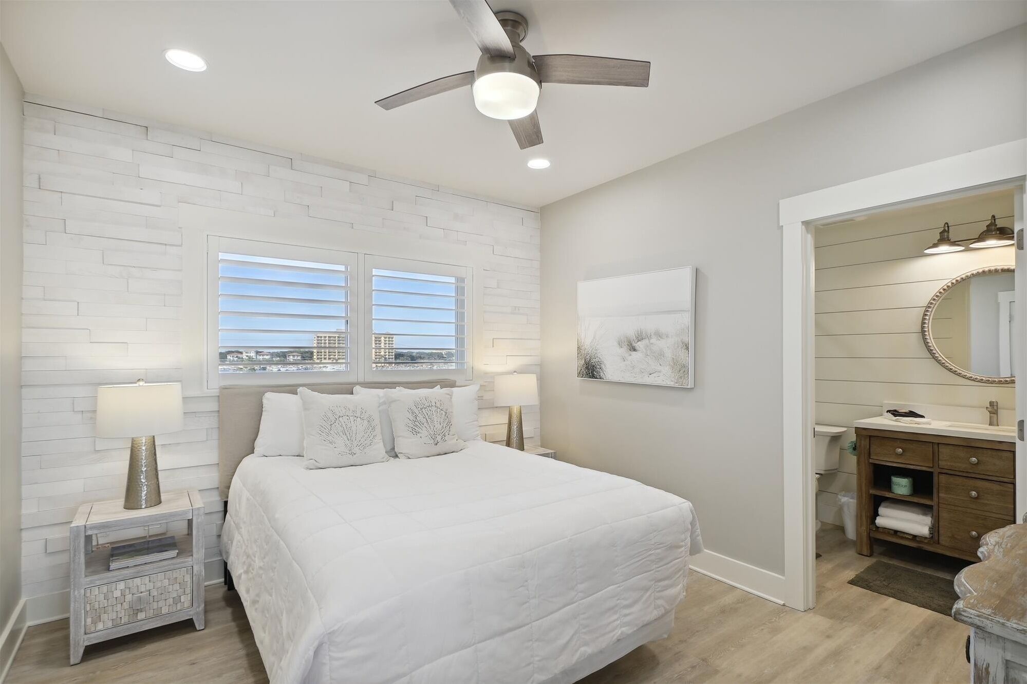 A bedroom with a bed, two bedside lamps, a ceiling fan, a window with blinds, a painting, and a bathroom with a vanity and mirror.