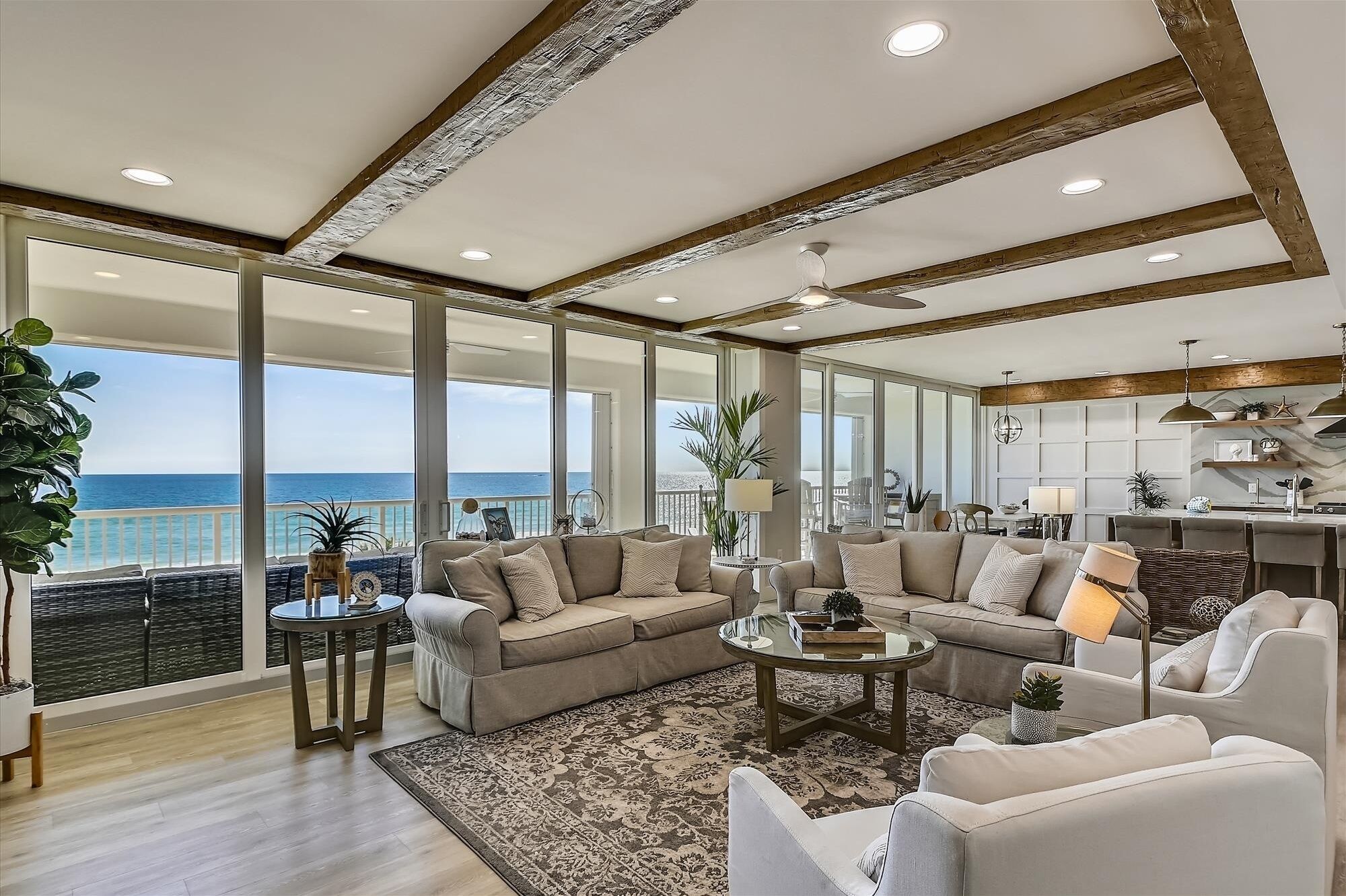 A spacious living room with a large window overlooking the sea, a cozy seating area with a coffee table, and a kitchen in the background.