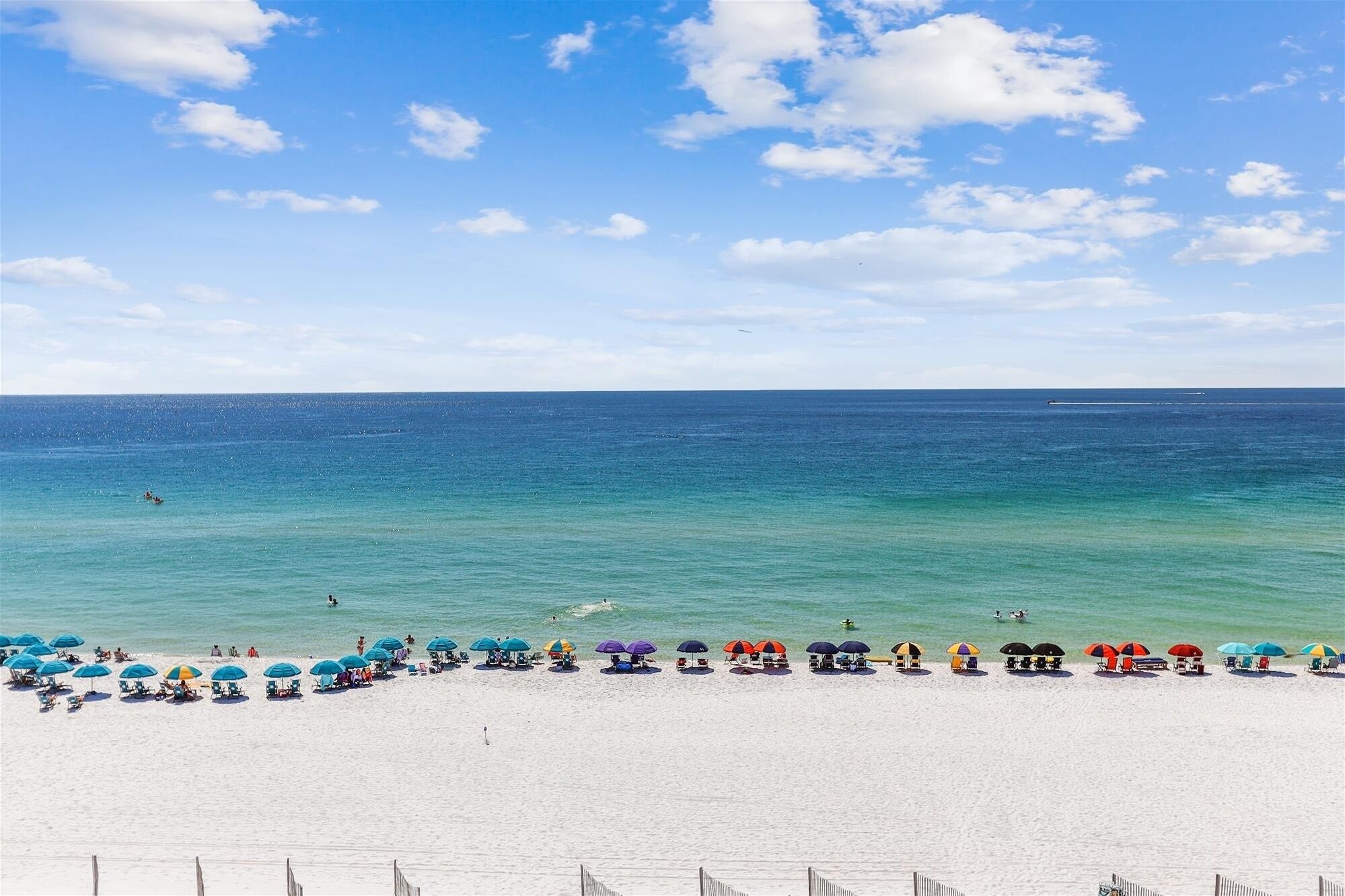 A beach with white sand and a clear blue sea. There are several umbrellas and lounge chairs along the shoreline.