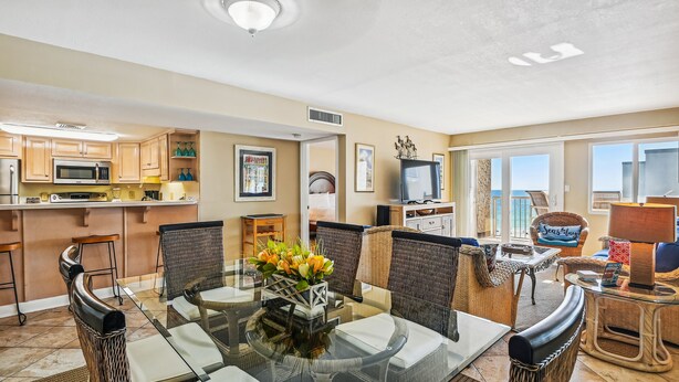 A modern kitchen with a microwave, stove, and bar area. There is a dining area with wicker chairs and a glass table. The room has a large window and a view of the city.