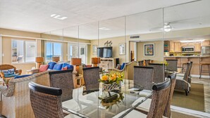 A spacious living area with a glass dining table, wicker furniture, and a kitchen in the background.
