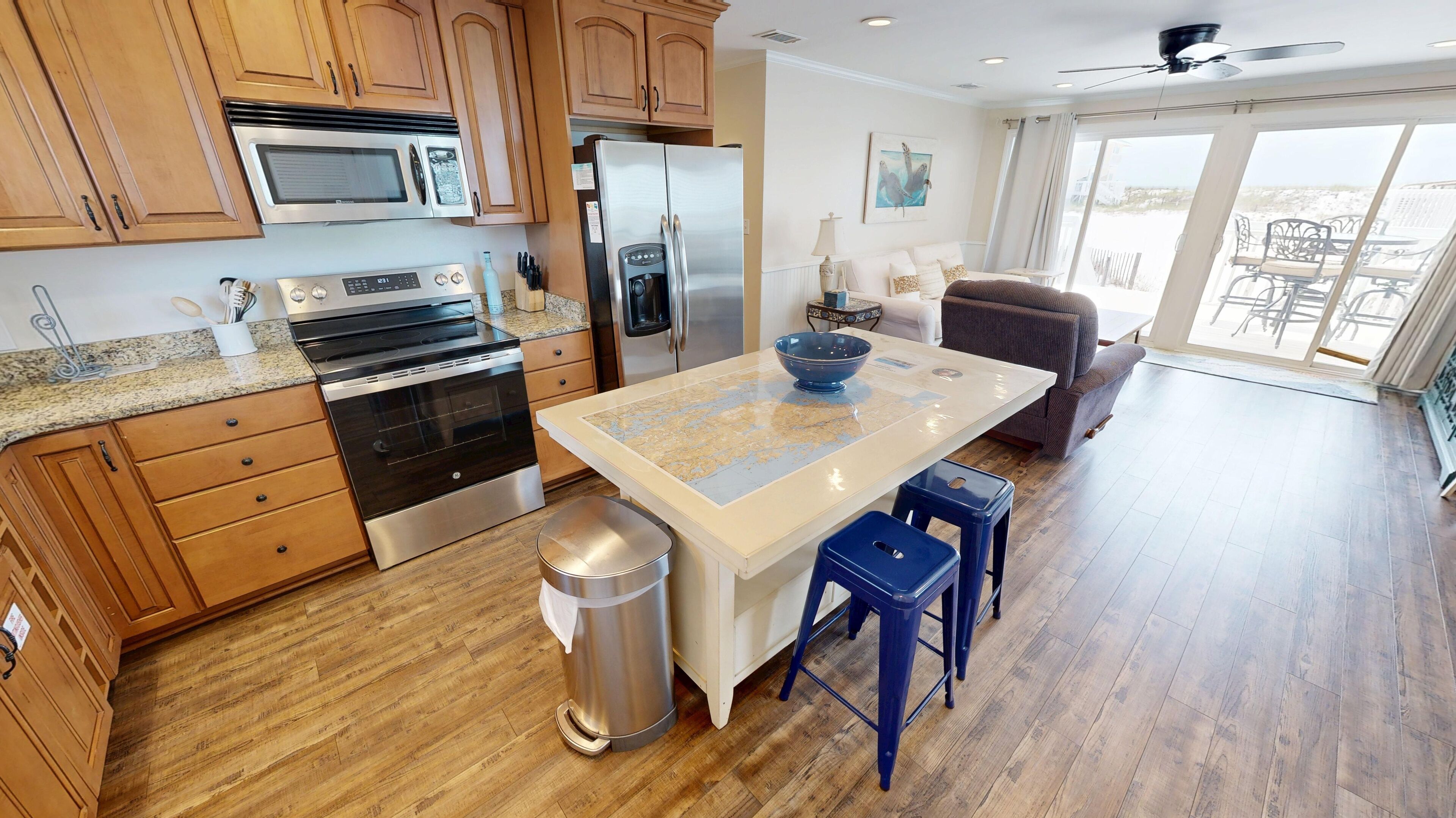 A modern kitchen with wooden cabinets, stainless steel appliances, a central island with stools, and a view of the outdoors through large windows.