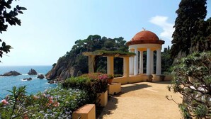 Un sentier côtier mène à un pavillon de style classique, surmonté d’un dôme, qui est entouré d’une végétation luxuriante et offre une vue sur la mer.