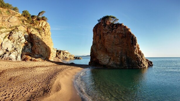 Une plage à l’eau claire, une grande formation rocheuse et un bâtiment situé sur la gauche.