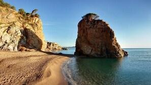 A beach with clear water, a large rock formation, and a building on the left. - Mas Ferran Adults Only (Pals)