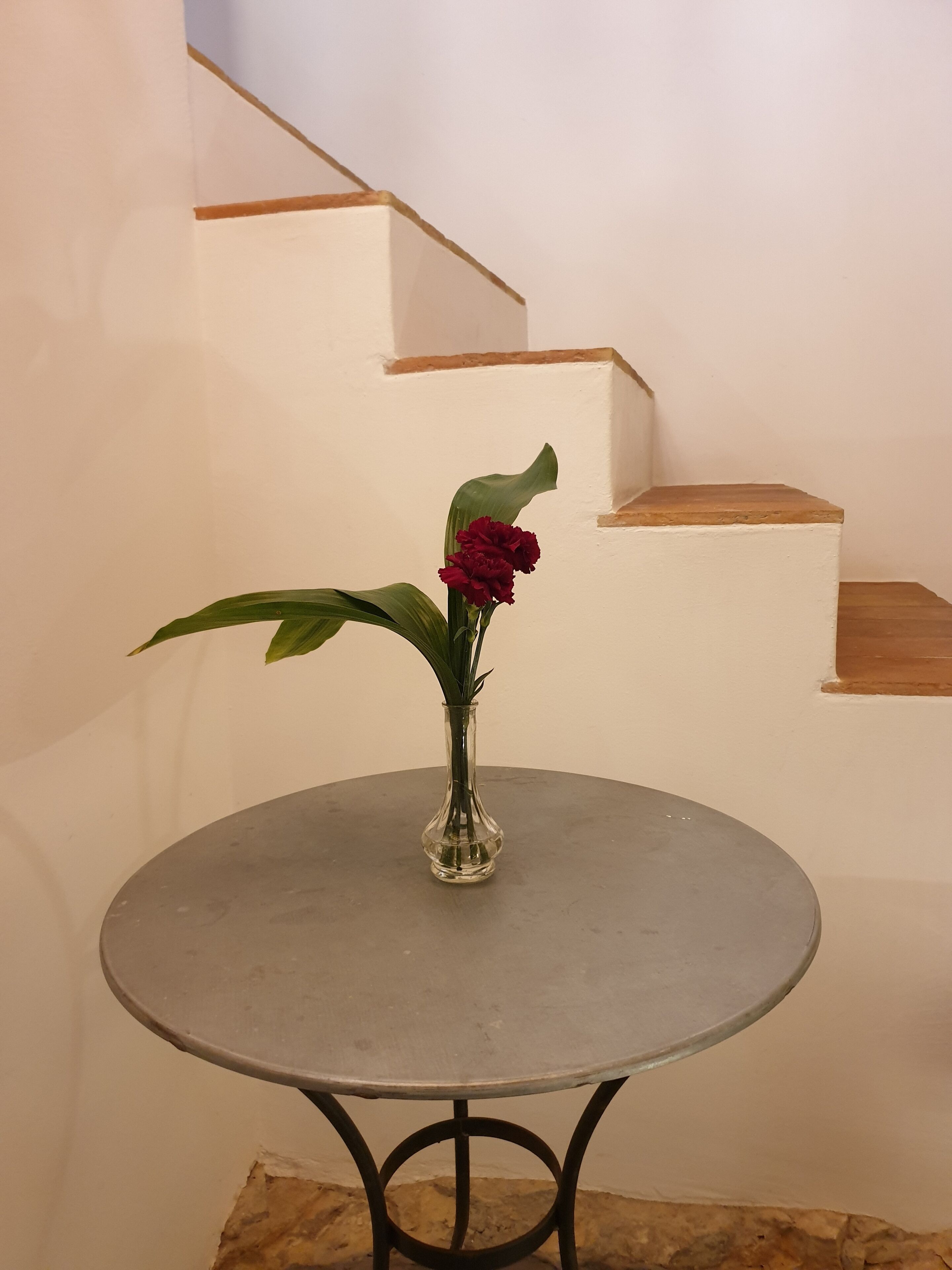A round table with a clear glass vase containing a single red flower and green leaves, positioned on a tiled floor with a staircase in the background.