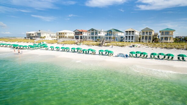 Een vakantiecomplex aan het strand met meerdere huizen, een zandstrand en groene parasols.