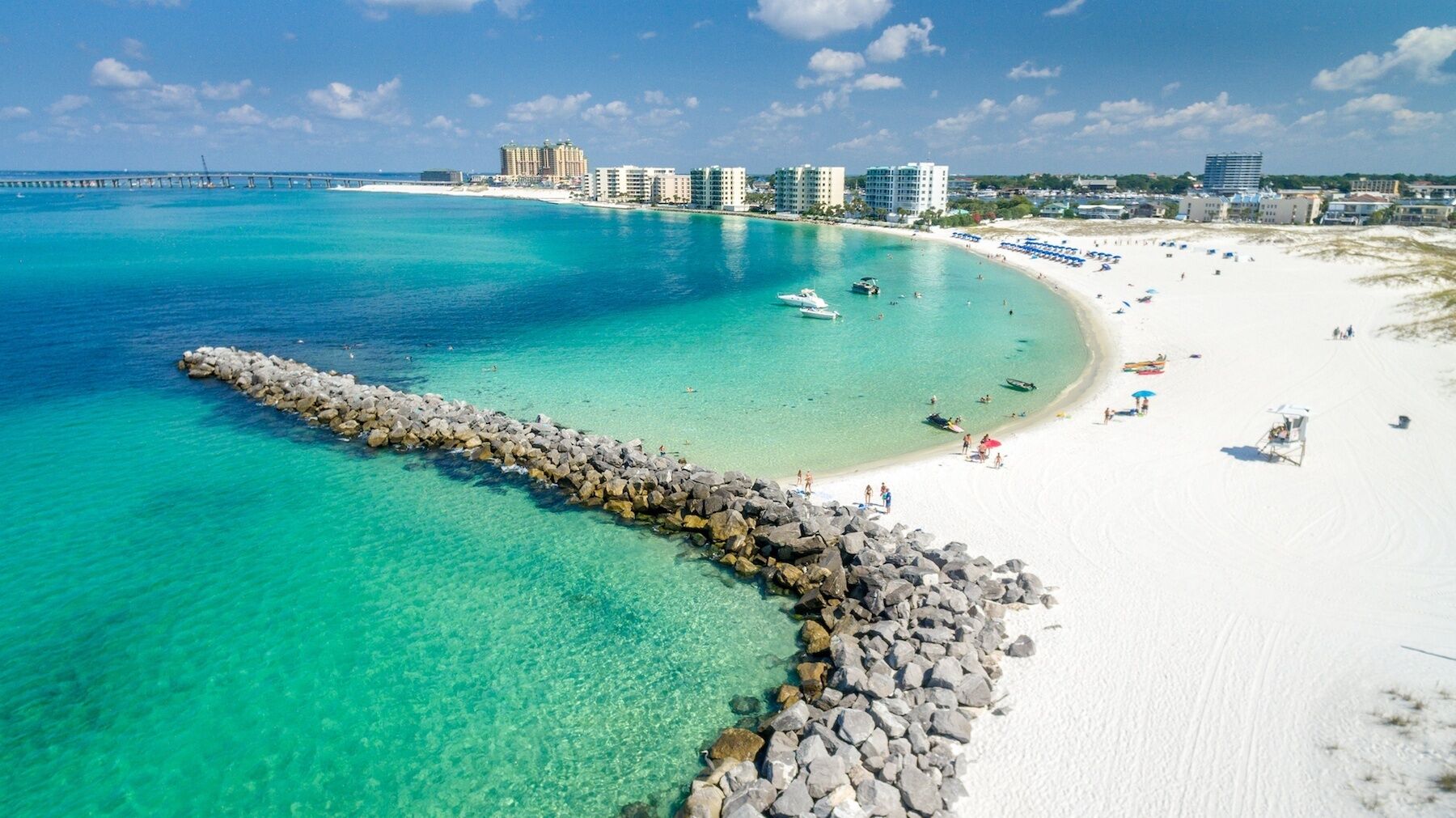 Une plage aux eaux turquoise et limpides, une digue rocheuse et une rangée de bâtiments situés en bord de mer.