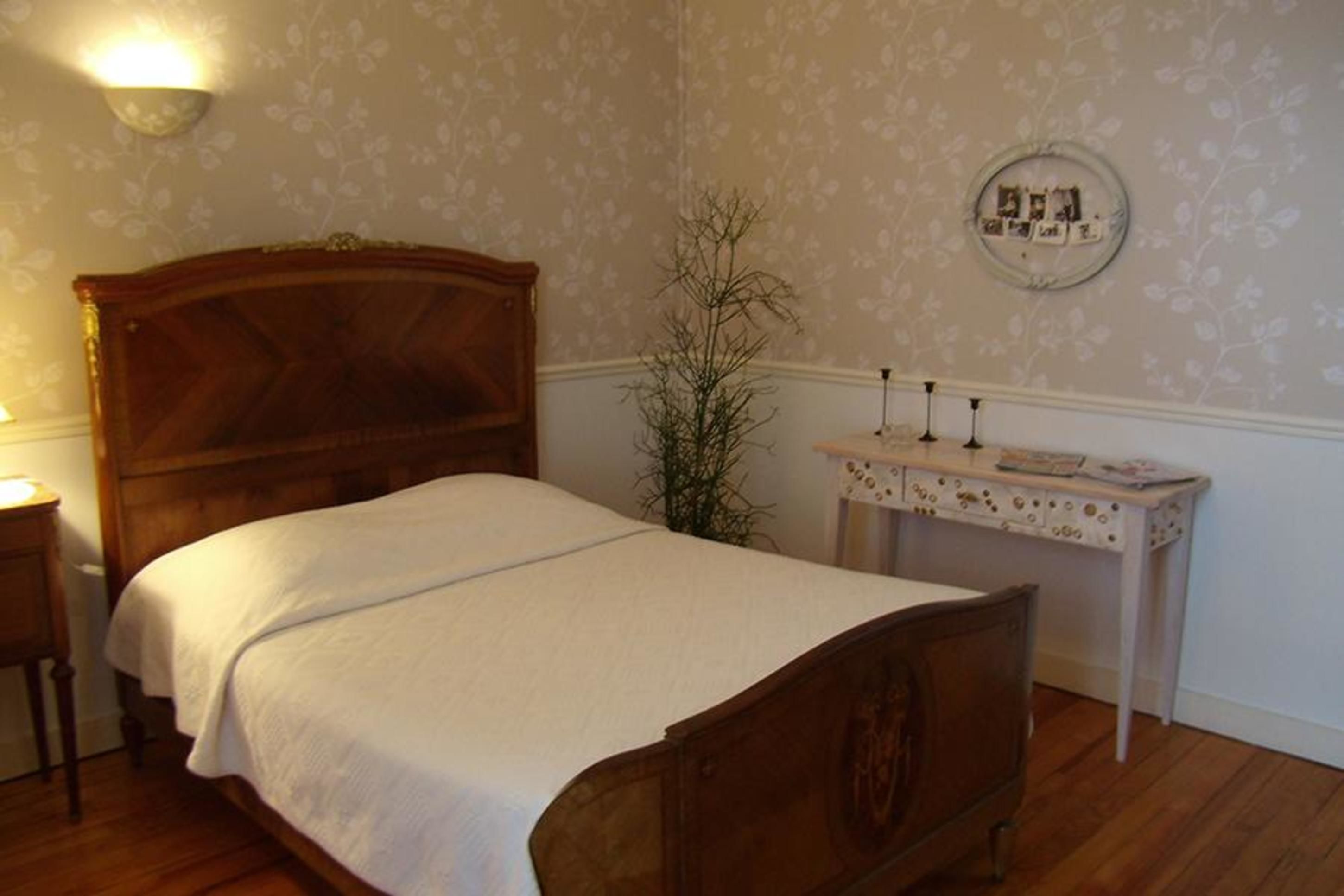 Un dormitorio con una cama de madera, una mesita de noche, una lámpara de pared, una pequeña mesa con un objeto decorativo y un reloj de pared.
