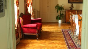 A room with a wooden herringbone floor, a red armchair, a dining table with chairs, a potted plant, and a door with a white frame. - Chambres d'hotes du Parc (Podensac)