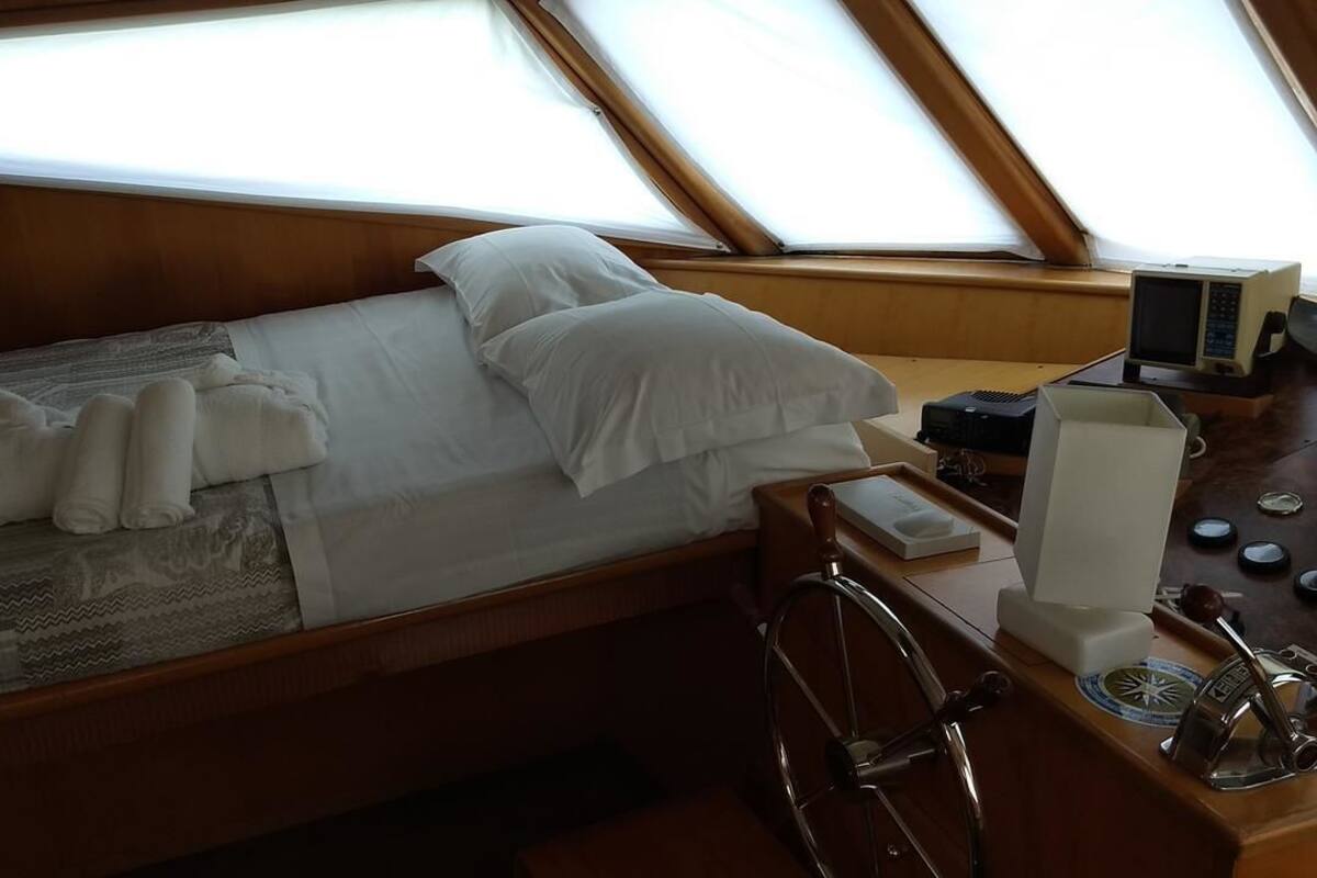 A small, cozy room with a single bed, a wooden headboard, a small table with a radio and a clock, and a window with a view of the sky.
