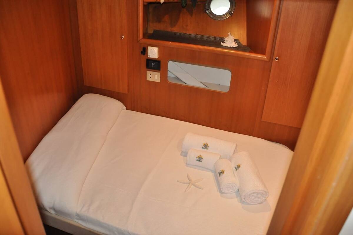 A small, neatly made bed with white linens and two rolled towels with a floral design.