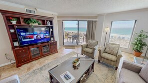 A living room with a flat-screen TV, a sofa, a coffee table, and a view of the beach through a sliding glass door.