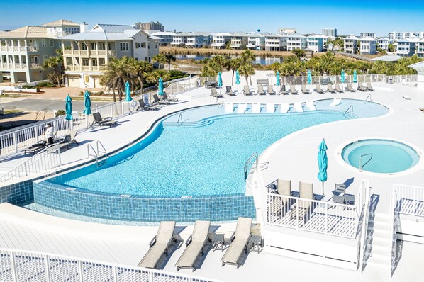Una piscina grande y curvada con sillas de descanso y sombrillas, rodeada por una terraza blanca y palmeras.