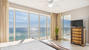 A bedroom with a large bed, a ceiling fan, a TV on a wooden cabinet, and a view of the ocean through large windows. - Oceania 907 3 Bedroom Condo by RedAwning (Destin)