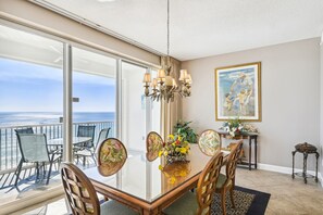 Ein Essbereich mit einem Glastisch, Stühlen und einem Kronleuchter. Ein Gemälde ist an der Wand und es gibt einen Blick auf das Meer durch große Fenster.