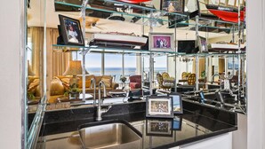 A modern kitchen with a black countertop, stainless steel sink, and glass shelves displaying various items. - Oceania 907 3 Bedroom Condo by RedAwning (Destin)