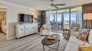 A spacious living room with a large flat-screen TV, a ceiling fan, a white sofa, a beige armchair, and a dining area with a view of the ocean.
