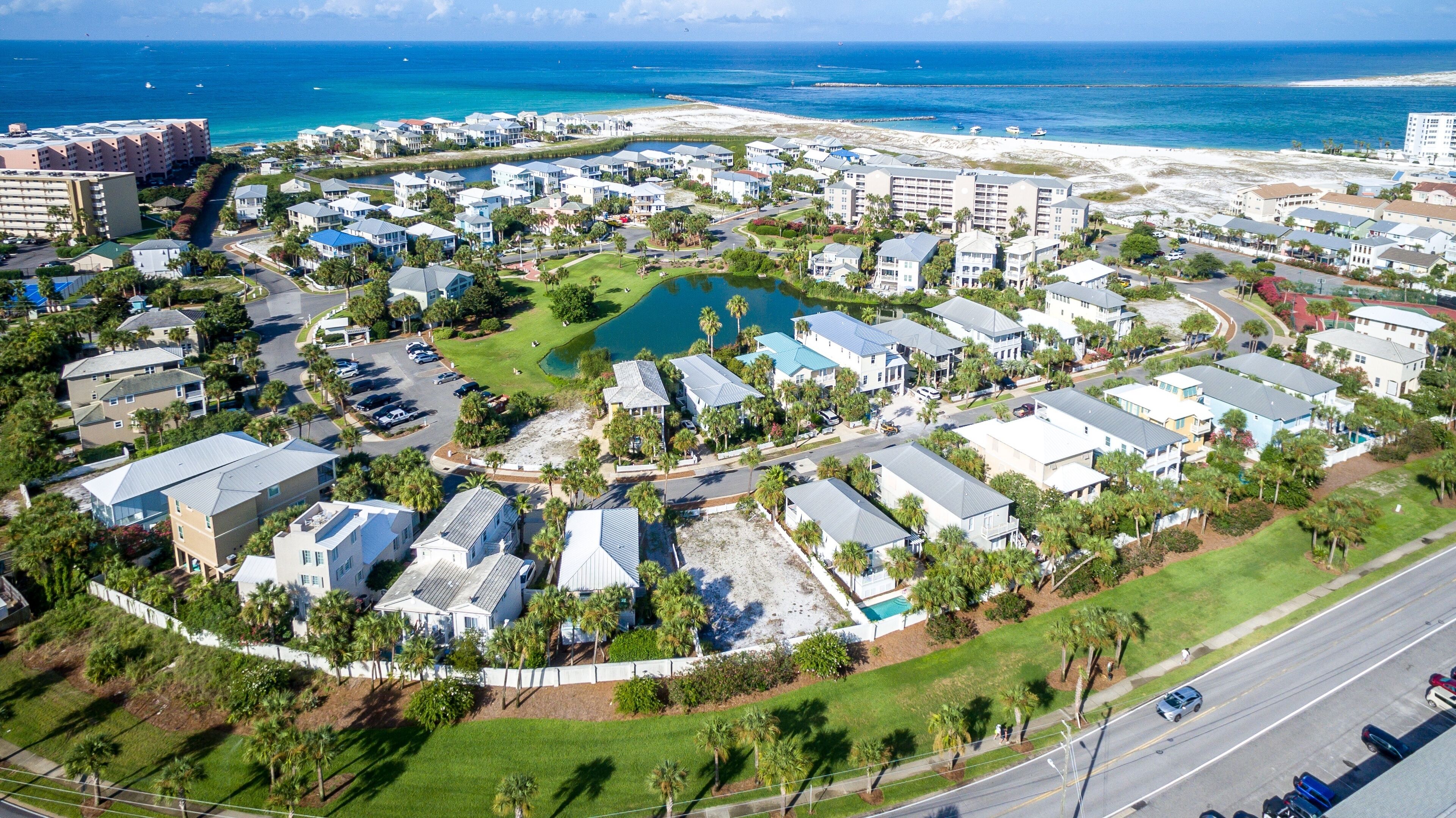 An aerial view of a coastal residential area with multiple buildings, a central green space, and a beach in the background.