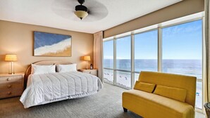 Hotelkamer met een groot bed, twee bedlampen, een plafondventilator, een groot raam met uitzicht op het strand en een gele fauteuil.